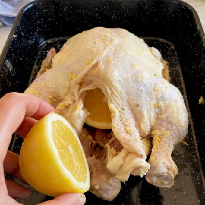 Lemon Garlic Butter Roast Chicken A raw whole chicken in a roasting pan with a hand holding a halved lemon, one half already placed inside the chicken cavity.