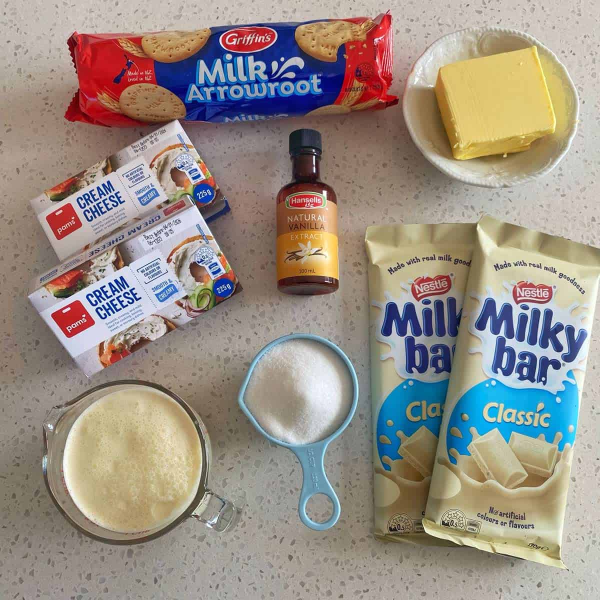 Milky Bar Cheesecake Ingredients for a dessert arranged on a countertop: Milk Arrowroot biscuits, cream cheese, vanilla extract, butter, Milky Bar chocolate, sugar, and a measuring cup of milk.