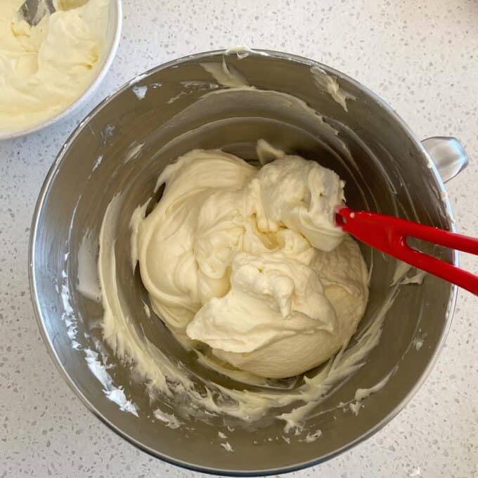 Milky Bar Cheesecake A mixing bowl filled with creamy, whipped frosting being stirred with a red spatula, placed on a light-colored countertop.