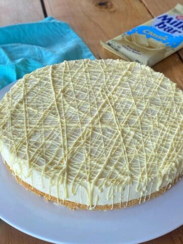 A white chocolate cheesecake with a biscuit base and white chocolate drizzle, displayed on a white plate with a blue cloth and a Milkybar chocolate bar in the background.