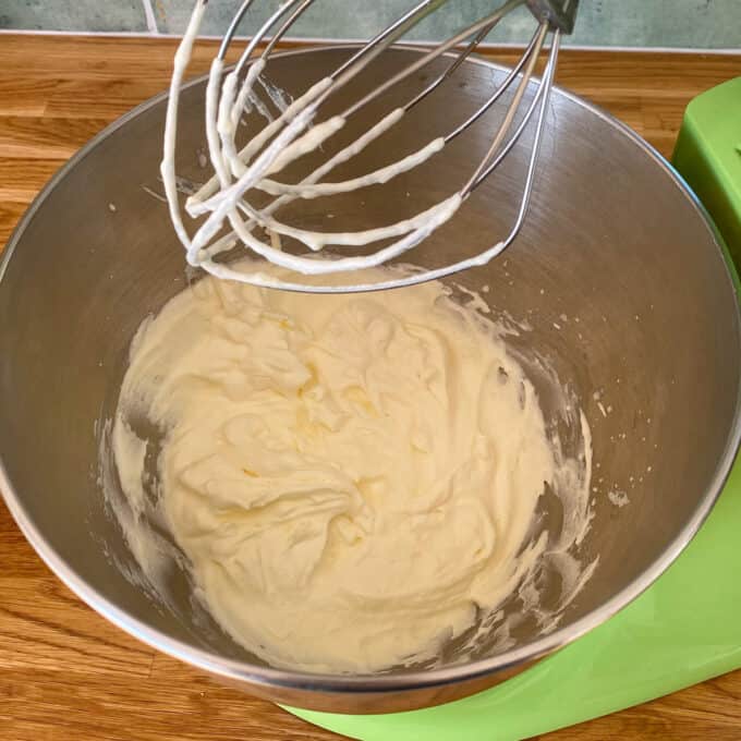 Milky Bar Cheesecake A metal mixing bowl with whipped cream mixture and a whisk attachment resting above it, placed on a wooden countertop.