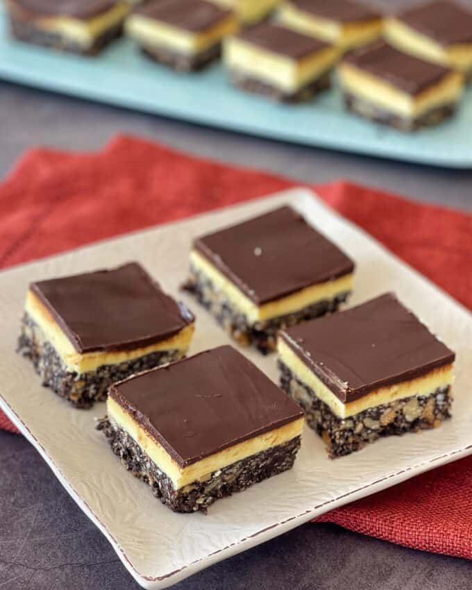 Nanaimo Slice Four Nanaimo bars with chocolate, custard, and coconut-crumb layers are arranged on a white plate, with more bars on a blue tray in the background.