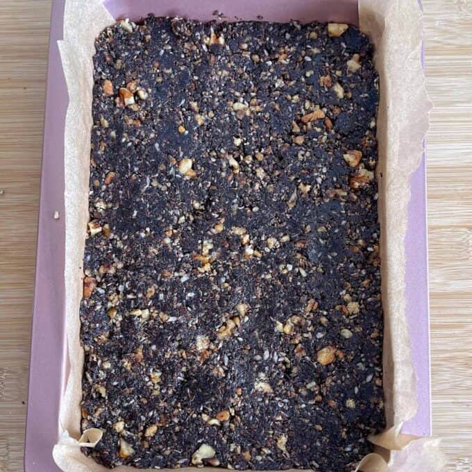 Nanaimo Slice A rectangular baking tray lined with parchment paper holds a pressed mixture of chopped nuts and dark, crumbly ingredients.