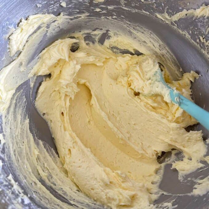 Nanaimo Slice A metal mixing bowl with creamy, pale yellow batter being mixed with a light blue spatula.