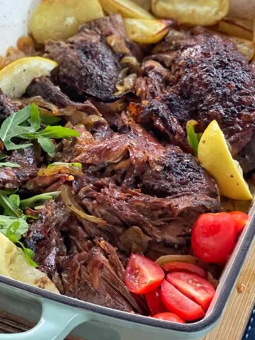 A roasted meat dish in a baking pan with potato wedges, arugula, lemon slices, and tomato pieces arranged on the side.