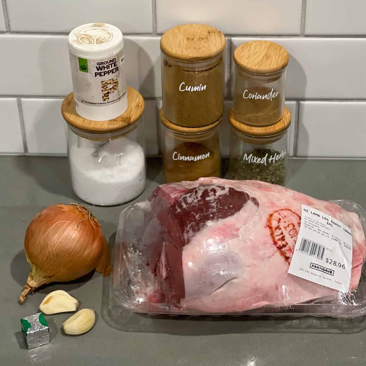Slow Cooker Lamb Shoulder A raw lamb leg roast, an onion, garlic cloves, a stock cube, salt, white pepper, cumin, coriander, cinnamon, and mixed herbs are arranged on a kitchen counter.