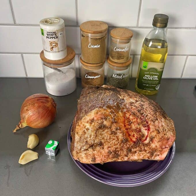 Slow Cooker Lamb Shoulder A raw seasoned roast sits on a plate beside an onion, garlic cloves, a stock cube, and jars of spices, salt, white pepper, and a bottle of olive oil on a kitchen counter.