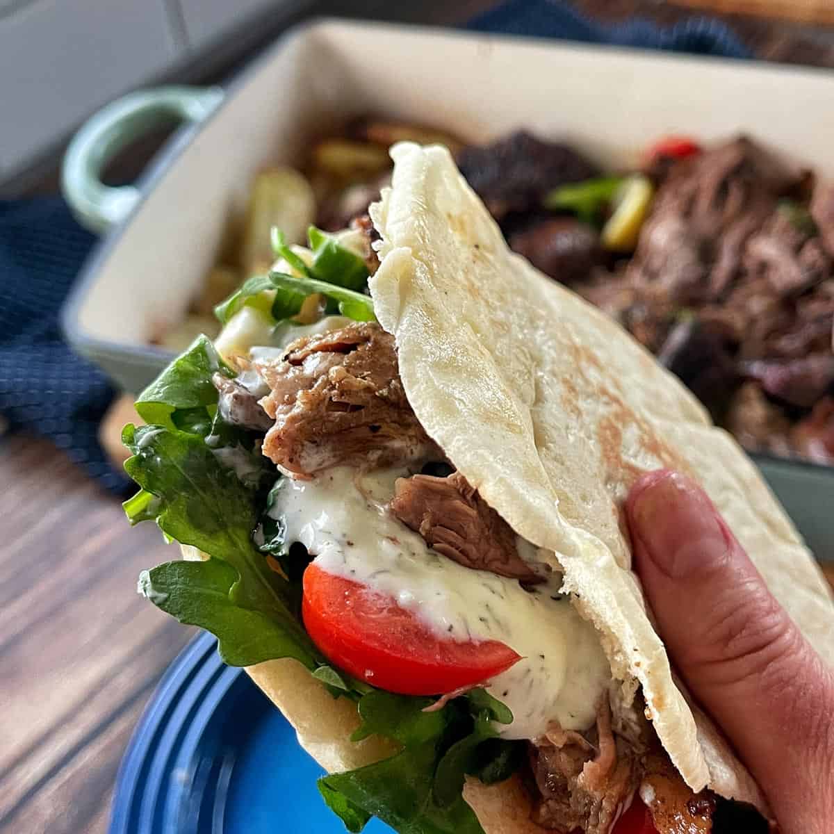 Slow Cooker Lamb Shoulder A hand holding a flatbread wrap filled with shredded meat, lettuce, tomato, and white sauce; a baking dish with more meat is in the background.