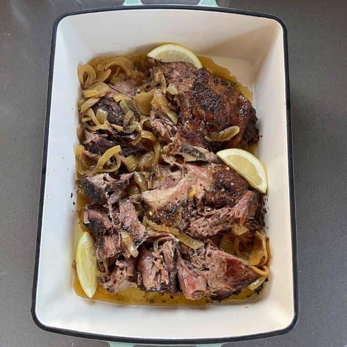 Slow Cooker Lamb Shoulder Cooked, shredded lamb shoulder with sliced onions and lemon wedges in a white rectangular roasting pan.