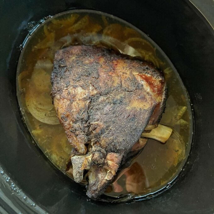 Slow Cooker Lamb Shoulder A cooked pork shoulder roast with seasoning rests in a slow cooker surrounded by broth and onions.
