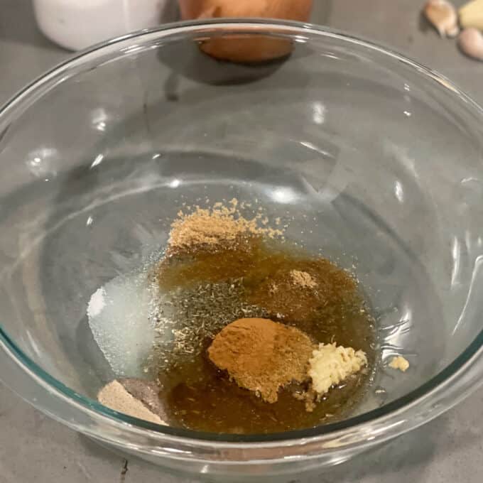 Slow Cooker Lamb Shoulder A glass bowl containing oil, ground spices, minced garlic, salt, and pepper on a gray countertop with ingredients in the background.