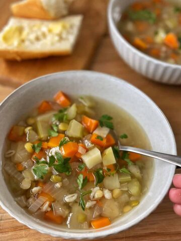 A bowl of vegetable soup with carrots, celery, and parsley, with a spoon in hand; sliced bread with butter on a wooden board in the background.