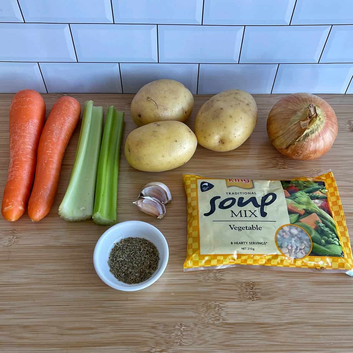 Vegetable Soup A wooden surface displays two carrots, two celery stalks, three potatoes, an onion, two garlic cloves, a bowl of dried herbs, and a packet of vegetable soup mix.