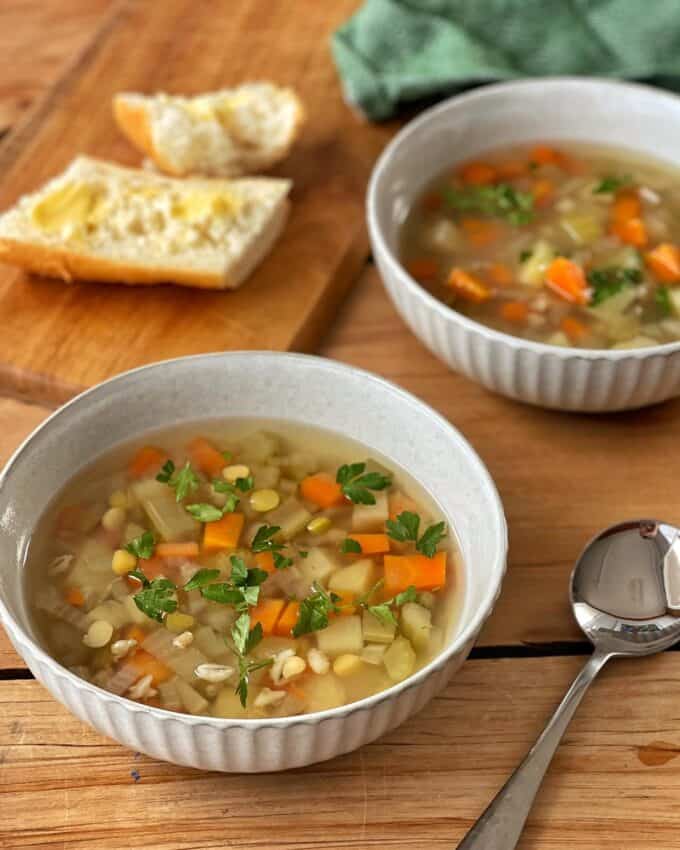 Vegetable Soup Two bowls of vegetable soup with carrots, celery, barley, and parsley on a wooden table, next to buttered bread slices and a spoon.