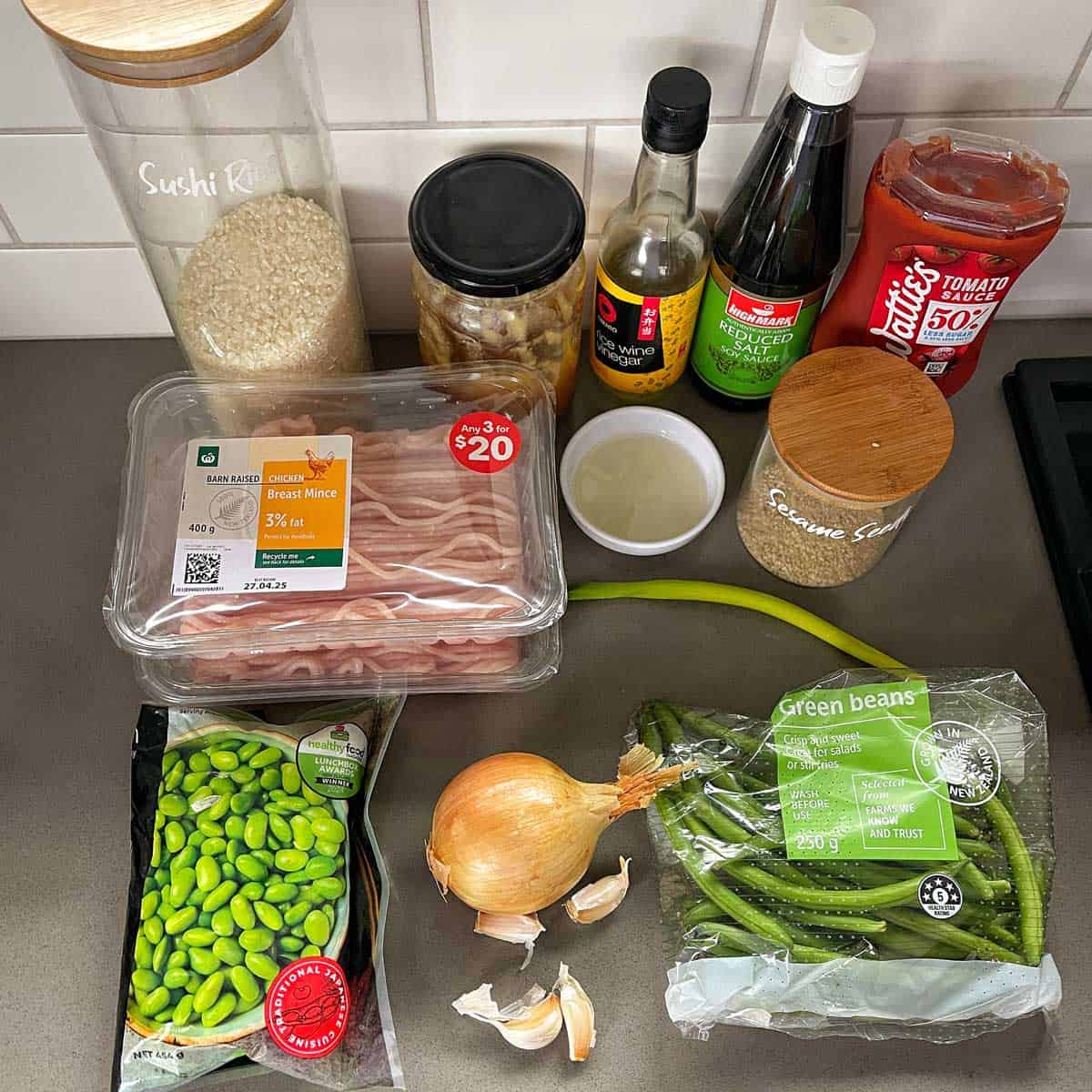 Sesame Chicken Rice Bowls A variety of ingredients on a kitchen counter, including rice, bacon, edamame, green beans, onion, garlic, sauces, tomato paste, and seasonings.