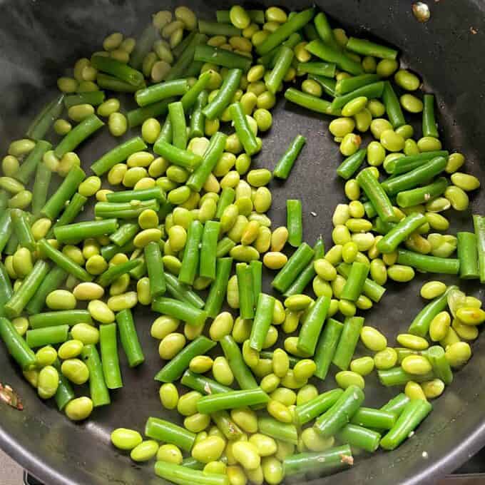 Sesame Chicken Rice Bowls Green beans and edamame are being sautéed in a black pan. Steam is visible on the left side, indicating they are being cooked.