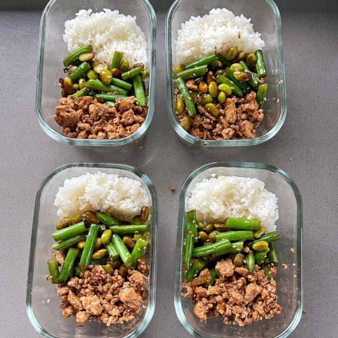 Sesame Chicken Rice Bowls Four glass meal prep containers each holding cooked white rice, green beans with edamame, and seasoned ground meat, arranged in a grid on a gray surface.