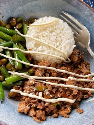 A bowl with white rice, sautéed green beans, minced meat, and mayonnaise drizzled on top, served with a fork.