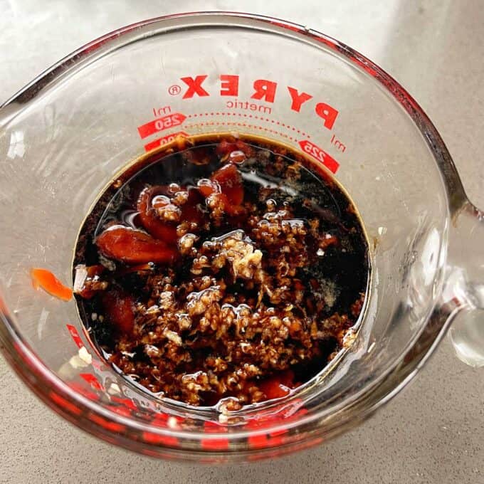 Sesame Chicken Rice Bowls A glass measuring cup filled with a mixture of dark liquid, minced garlic, and a red sauce on a kitchen countertop.