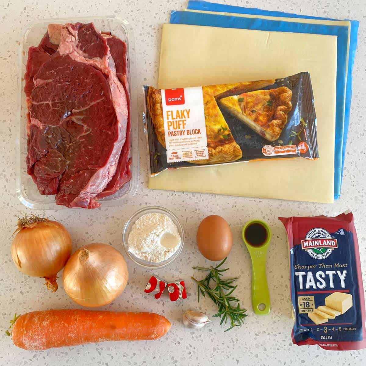 Steak and Cheese Pie Ingredients on a countertop include beef, puff pastry, onions, carrot, cheese, egg, flour, butter, herbs, chilies, and soy sauce.
