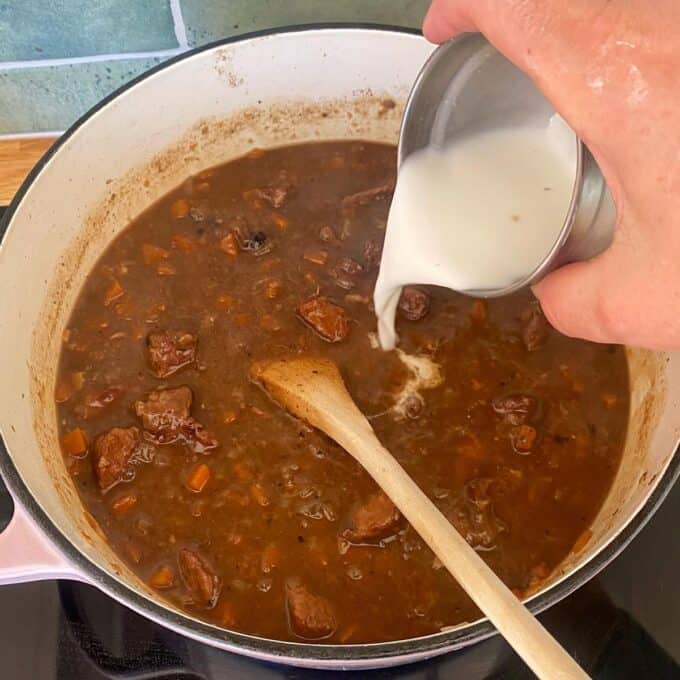 Steak and Cheese Pie A hand pours a white liquid, likely a slurry or cream, into a pot of simmering brown stew with chunks of meat and vegetables, while a wooden spoon rests inside the pot.