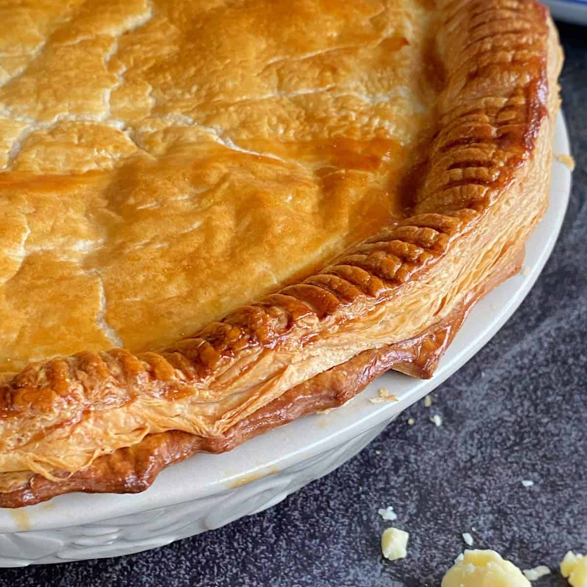 Steak and Cheese Pie A golden-brown baked pie with a flaky crust sits in a white ceramic pie dish on a dark textured surface.