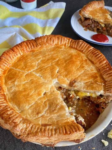 A baked meat pie with a golden crust sits in a dish, with a large slice missing; the slice is served on a plate nearby with some ketchup.