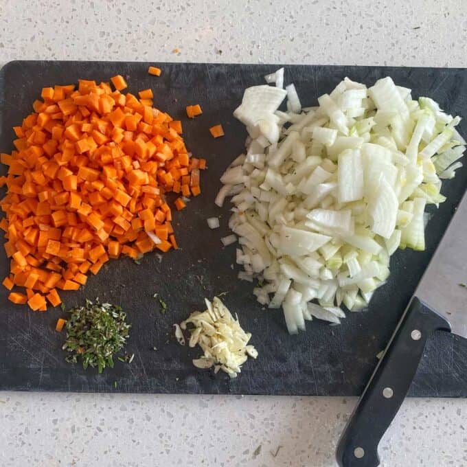 Steak and Cheese Pie Diced carrots, chopped onions, minced garlic, and chopped herbs on a black cutting board next to a chef’s knife.