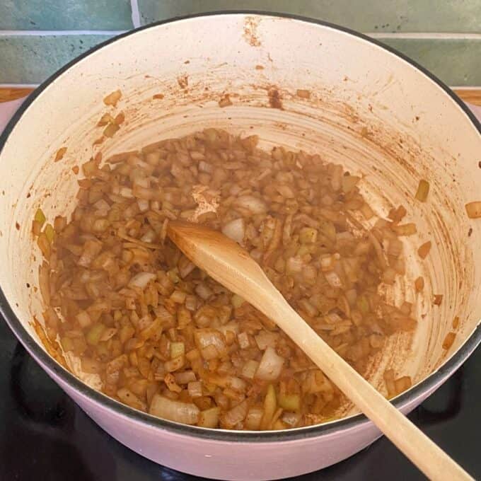 Steak and Cheese Pie Chopped onions being sautéed with spices in a large white pot, stirred with a wooden spoon on a stovetop.