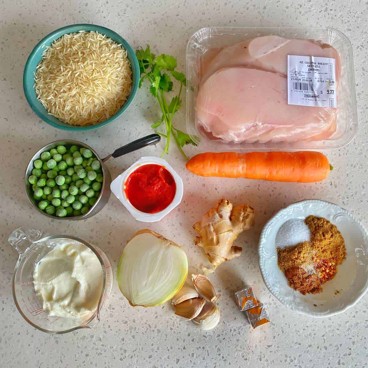 Chicken Tikka Tray Bake A countertop displays raw chicken breasts, a bowl of rice, fresh cilantro, a carrot, frozen peas, tomato sauce, yogurt, onion, ginger, garlic, bouillon cubes, and spices.