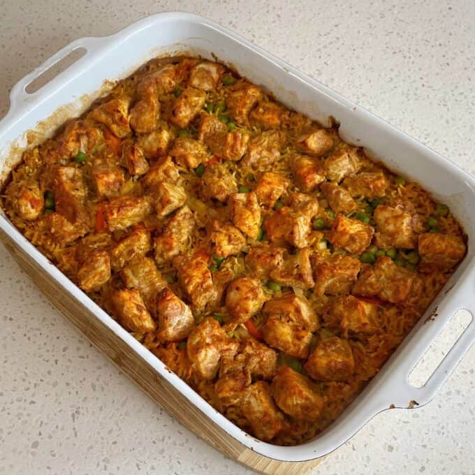 Chicken Tikka Tray Bake A white baking dish filled with baked rice, peas, carrots, and seasoned chunks of chicken on a speckled countertop.