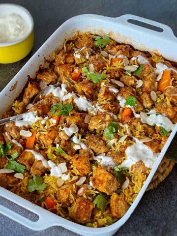 A white baking dish filled with baked biryani topped with cilantro, sliced almonds, and a yogurt drizzle; a bowl of yogurt is beside it.