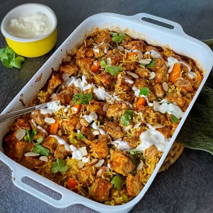 Chicken Tikka Tray Bake A white baking dish filled with baked biryani topped with cilantro, sliced almonds, and a yogurt drizzle; a bowl of yogurt is beside it.