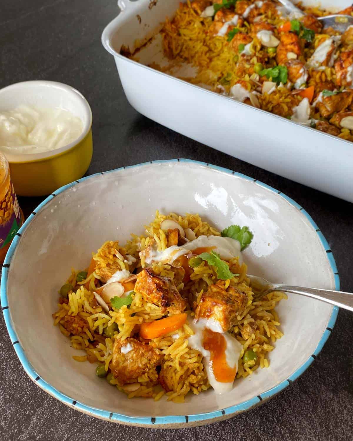 Chicken Tikka Tray Bake A bowl of biryani with pieces of chicken, vegetables, and fresh herbs, topped with yogurt, with a baking dish of more biryani and a bowl of yogurt in the background.
