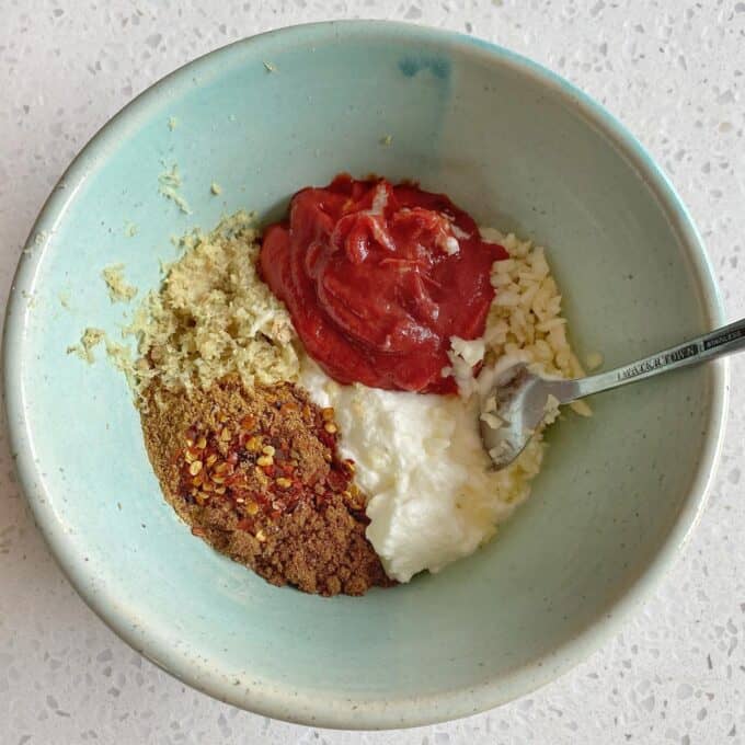 Chicken Tikka Tray Bake A bowl containing grated ginger, tomato paste, minced garlic, yogurt, ground spices, and red pepper flakes, with a spoon resting inside.