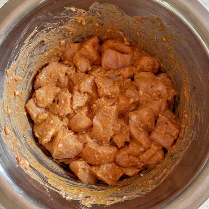Chicken Tikka Tray Bake Cubed pieces of raw chicken marinating in a seasoned reddish sauce inside a stainless steel mixing bowl.