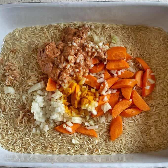 Chicken Tikka Tray Bake Uncooked rice in a baking dish topped with chopped onions, sliced carrots, turmeric, and a dollop of a brownish sauce or paste.