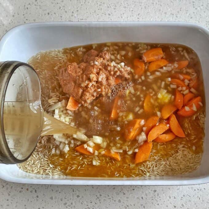 Chicken Tikka Tray Bake A baking dish with uncooked rice, chopped onions, carrots, and sauce, as broth is being poured over the mixture on a speckled countertop.