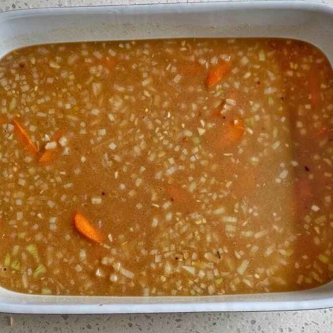 Chicken Tikka Tray Bake A rectangular white baking dish filled with a broth containing chopped onions, carrot slices, and spices on a speckled countertop.