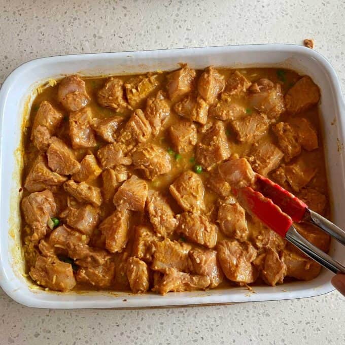 Chicken Tikka Tray Bake Marinated chunks of raw chicken arranged in a white baking dish, with red tongs holding one piece over the dish on a light countertop.