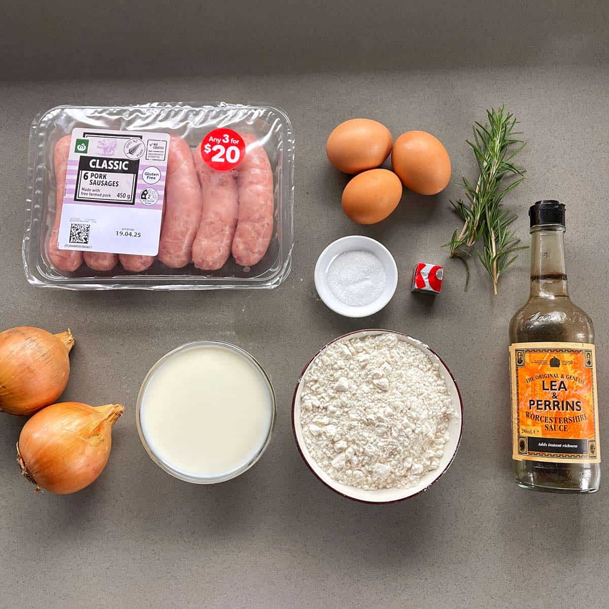 Toad in the Hole A countertop displays raw sausages, three eggs, rosemary, salt, two onions, milk, flour, Worcestershire sauce, and a red-capped stock cube.