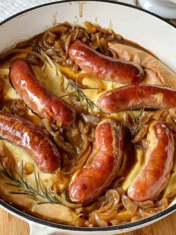 A baked dish with browned sausages, caramelized onions, and sprigs of rosemary on a golden batter in a white casserole dish.