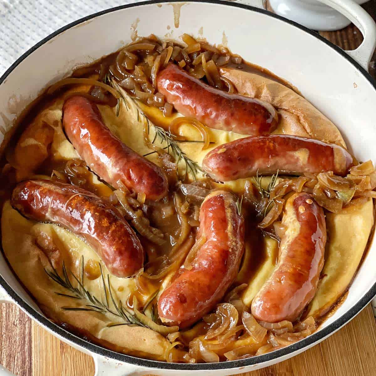 Toad in the Hole A baked dish with browned sausages, caramelized onions, and sprigs of rosemary on a golden batter in a white casserole dish.