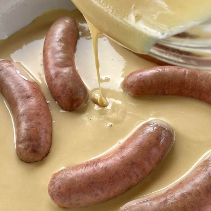 Toad in the Hole Uncooked sausages in a baking dish being covered with a thick batter, possibly for toad in the hole preparation.