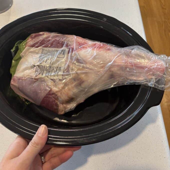 Slow Cooker Lamb Shoulder A raw Slow Cooker Spiced Lamb Shoulder in plastic packaging sits inside a black slow cooker on a kitchen countertop.
