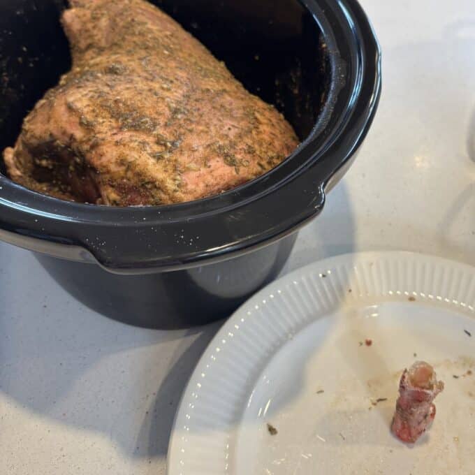 Slow Cooker Lamb Shoulder Seasoned Slow Cooker Spiced Lamb Shoulder sits in the slow cooker, while a small meat-covered bone rests on a white plate beside it on the countertop.