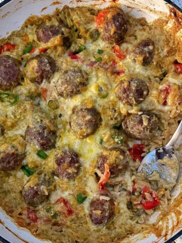 A baked dish inspired by Creamy Pesto Orzo with Meatballs, featuring meatballs, rice, melted cheese, chopped tomatoes, and green onions in a white pan with a serving spoon inside.