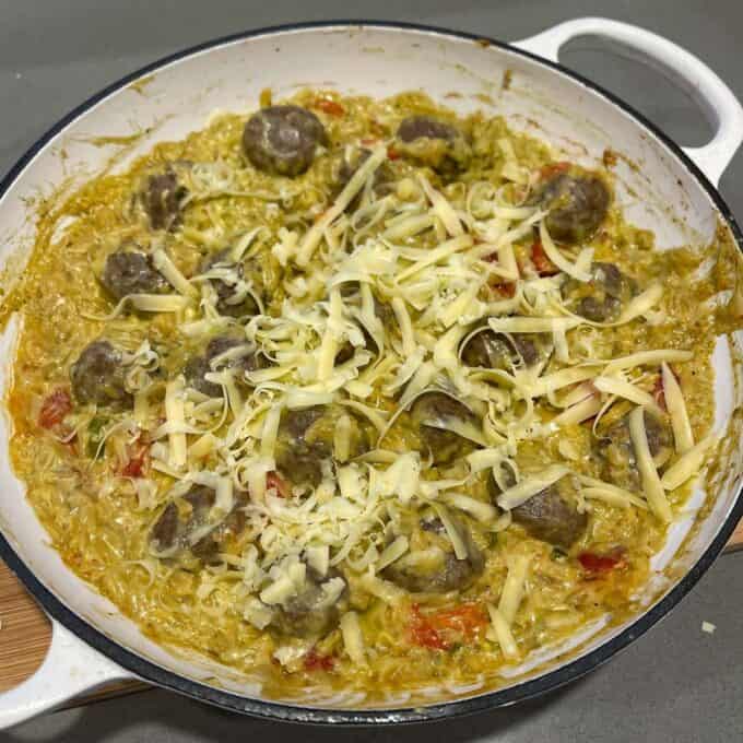 Creamy Pesto Orzo and Meatballs A white skillet filled with creamy pasta, meatballs, chopped tomatoes, and topped with shredded cheese, resting on a gray countertop.