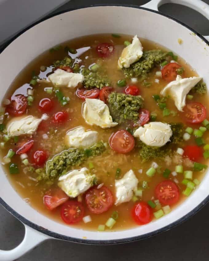 Creamy Pesto Orzo and Meatballs A pot containing broth, halved cherry tomatoes, green onions, dollops of cream cheese, and spoonfuls of pesto.