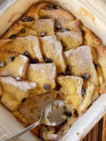 A baked bread and butter pudding with raisins in a square dish, topped with powdered sugar. A serving spoon rests inside, indicating a portion has been taken out.